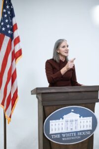 A woman speaks confidently at a podium with the American flag and White House insignia.