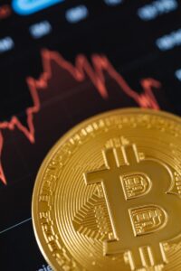Close-up of a gold Bitcoin coin with a fluctuating stock market graph in the background.