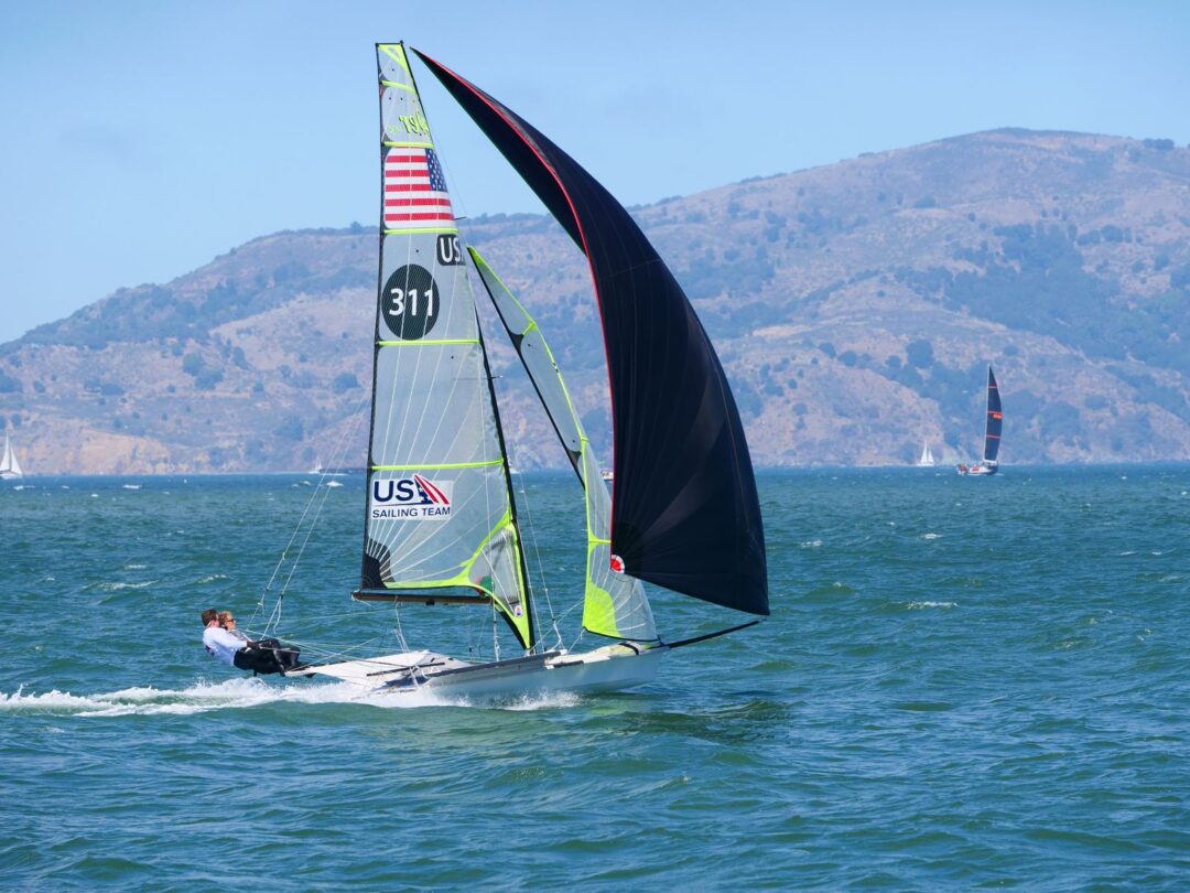 Dynamic sailboat race on a sunny day in San Francisco Bay against a mountainous backdrop.